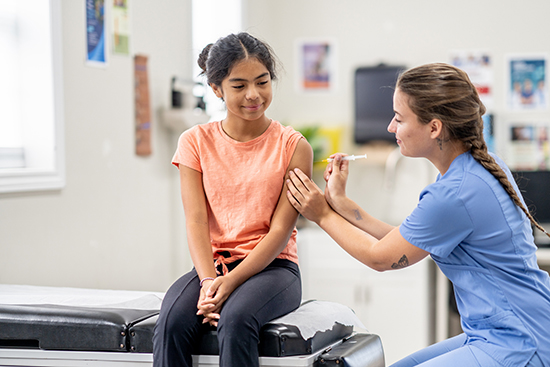 Teen girl getting vaccine in arm from woman in blue scrubs.