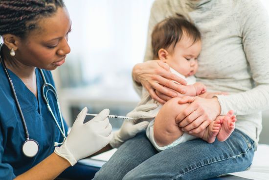 A healthcare professional prepares to administer a vaccine to a baby sitting on a caregiver's lap in a clinic.