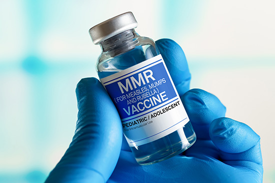 A gloved hand holds up a vial labeled "MMR VACCINE"