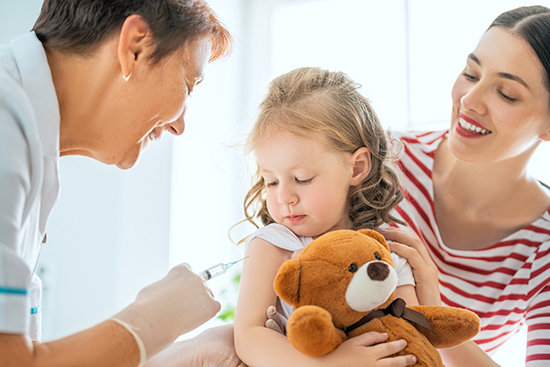 Young girl holding teddy bear getting vaccine in arm.